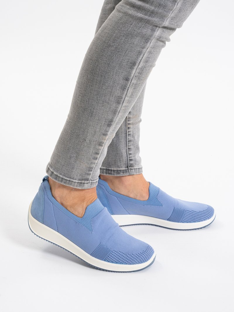 Person trägt hellblaue Slip-on-Sneaker mit weißen Sohlen und graue eng anliegende Jeans, steht vor einem weißen Hintergrund.
