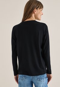 Svart långärmad topp med rund hals, mjuk textur och normal passform, tillsammans med ljusblå denimjeans. Bakifrån.