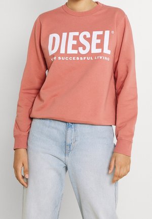 Persoon met een roze Diesel-trui met lange mouwen en witte tekst, en lichtblauwe hooggetailleerde spijkerbroek, zicht van taille tot borst.