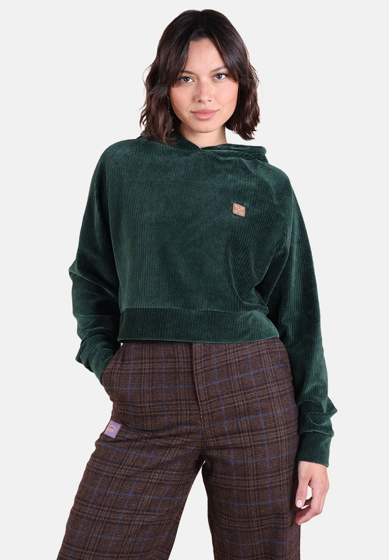 Haut à capuche en velours côtelé vert avec un design court et une petite étiquette logo. Le modèle porte un pantalon à carreaux marron avec des lignes bleu accent.