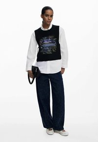 Gilet noir en maille avec un motif à paillettes, chemise blanche à col en dessous, associé à un pantalon large paillettes bleu marine et des chaussures claires.