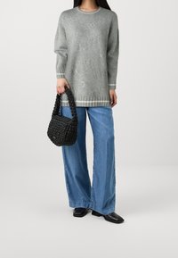 Suéter gris extragrande con puños acanalados, combinado con pantalones vaqueros de pierna ancha en azul claro. Sosteniendo un bolso tejido negro. Zapatos negros de punta cuadrada.