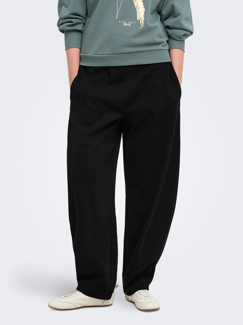 Personne portant un pantalon noir ample avec les mains dans les poches, associé à des baskets blanches et un sweat-shirt vert.
