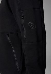 Sort sweatshirt med lynlåslommer, struktureret stof og et mat logo-patch på ærmet. Har et moderne, tætsiddende design.