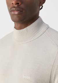 Suéter de cuello alto beige claro en punto fino con cuello acanalado. Presenta un sutil logo "BOSS" bordado en blanco en el pecho. Textura suave.