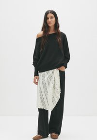 Zwarte oversized trui, witte kant rok met een diagonaal patroon, donkere jeans en bruin vlakke schoenen met versieringen. Eenvoudige, gestructureerde outfit.