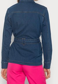 Jeansjacka med krage och bälte i midjan, med kontrasterande stygn och en mörkblå färg. Baksidan har en enkel design utan synliga fickor.