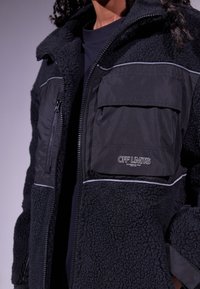 Giacca in fleece nera con una finitura morbida e testurizzata, tasche con zip e a pattina, e piping a contrasto lungo i bordi. Logo "OFF LIMITS" visibile.