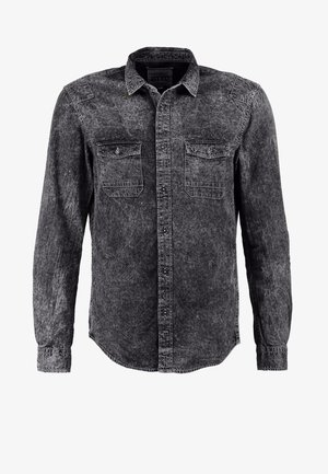 Mann mit tätowierten Armen, der ein graues Denimhemd, beige Hosen, graue Sneakers trägt und einen schwarzen Rucksack über einer Schulter trägt.