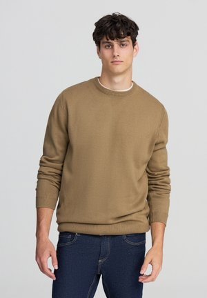 Maglione marrone lavorato a maglia con scollatura tonda, polsini e orlo a costine. Indossato sopra jeans blu scuro, il tessuto testurizzato aggiunge profondità.