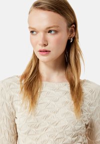 Cremefarvet top med tekstureret stof, der har et mønster af hævede, sammenvævede linjer. Modellen har lige, blond hår og sølvøreringe.