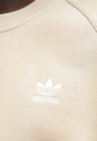 Béžová mikina Adidas s rebrovaným okrúhlym výstrihom, s bielym vyšívaným logom a hladkou, textúrovanou látkou.