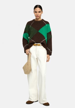 Kobieta ubrana w brązowo-zielony sweter w romby, białe szerokie spodnie z żółtym paskiem, brązowe buty, trzymająca beżową torbę obok siebie.