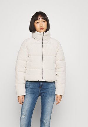 Hele beež puffer-jakk kõrge krae, tikandit tekstuur ja esine zipper; kantud siniste rebitud teksade peal.
