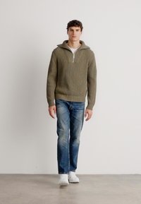 Maglione a coste verde oliva con colletto a mezza zip, indossato sopra una camicia bianca e abbinato a jeans blu sbiaditi e sneakers bianche.