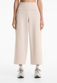 Beige geribde wijde broek met een hoge taille. Zachte textuur, geen zichtbare patronen. Model draagt lichtgrijze sneakers en witte crew-sokken.