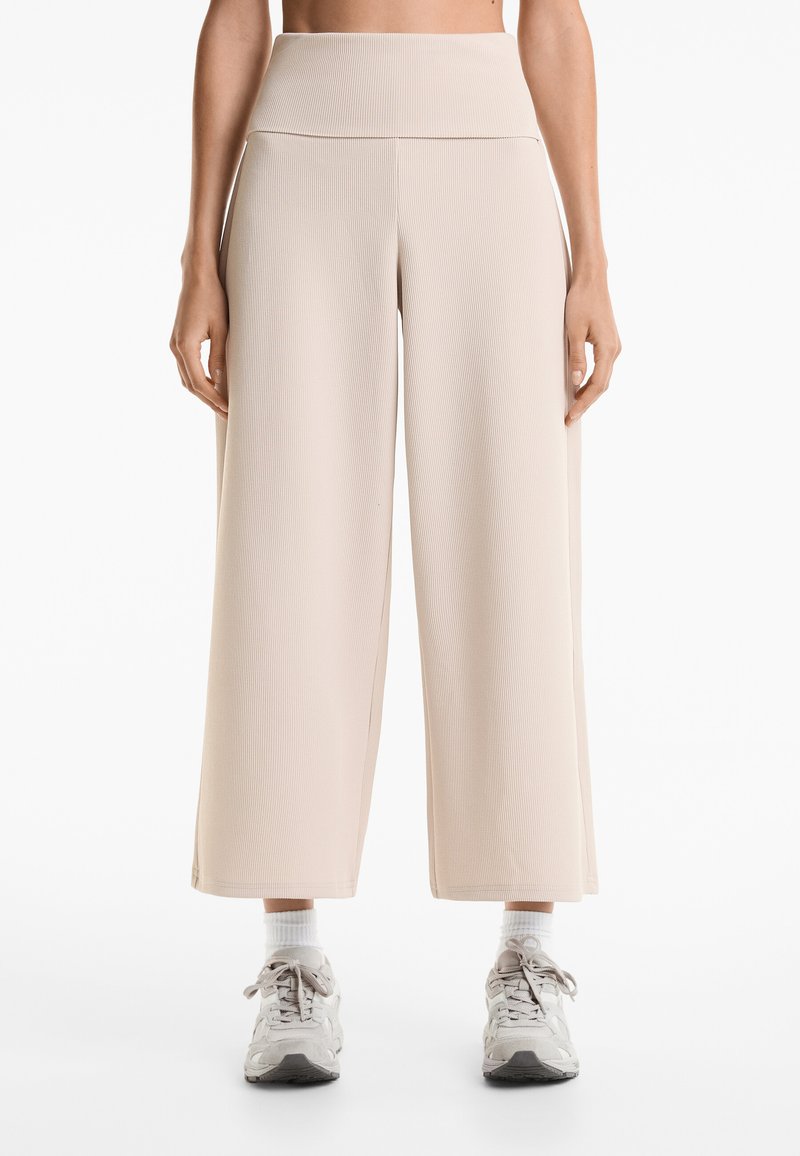 Beige geribde wijde broek met een hoge taille. Zachte textuur, geen zichtbare patronen. Model draagt lichtgrijze sneakers en witte crew-sokken.