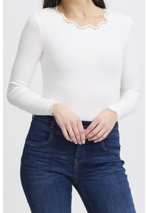 Femme portant un haut blanc à manches longues avec une bordure en dentelle au niveau de l'encolure et des poignets, associée à un jean bleu foncé, mains jointes devant.