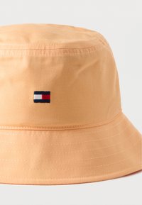 Πορτοκαλί καπέλο τύπου κάπα, κατασκευασμένο από μαλακό ύφασμα, με μικρό κεντημένο λογότυπο σε κόκκινο, λευκό και μπλε στο μπροστινό μέρος. Ομαλή υφή, στρογγυλό σχήμα.