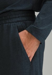 Tmavošedé športové nohavice s textúrovaným povrchom, elastickým pásom a bočnou vreckom. Ruka je vložená vo vrecku, čo ukazuje detail rukávu.