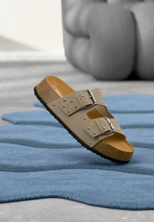 Beige sandaal met kurken zool, voorzien van twee met studs versierde banden en zilveren gespen, rustend op een blauw gestructureerd tapijt, met wazig grijs meubilair op de achtergrond.