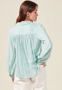 Blusa leggera a maniche lunghe di colore verde menta con strisce verticali bianche. Presenta dettagli plissettati sul retro e polsini con bottoni.