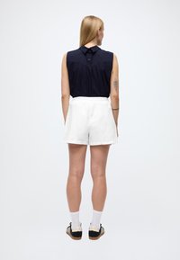 Pikkade blondide juustega naine, riietatud varrukateta tumesinisesse pluusi, valgetesse lühikutesse pükstesse, valgetesse sokkidesse ja mustadesse tossudesse, seisab näoga eemale tavalise tausta ees.