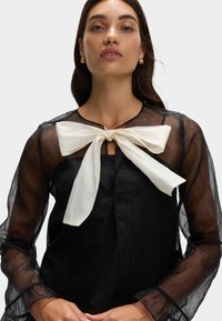Blusa negra transparente con un gran lazo blanco en el escote, mangas largas con bordes rizados y una tela texturizada.