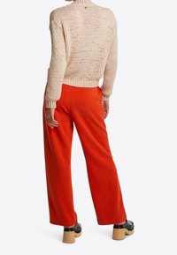 Pletený béžový svetr s žebrovanými manžetami a lemem, doplněný širokými oranžovými kalhotami. Černé dřeváky s dřevěnými podpatky dotvářejí vzhled.