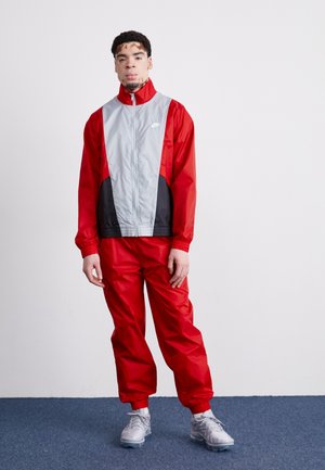 Red and gray windbreaker jacket with black accents, zip closure, and elastic cuffs, paired with matching red pants. Gray sneakers complete the outfit.
