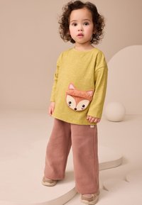 Maglietta a maniche lunghe verde chiaro con tasca applicata a forma di volpe e dettagli rosa, abbinata a pantaloni marroni larghi. Tessuto liscio, design casual.