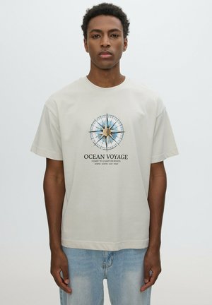 Joven con cabello corto y rizado que lleva una camiseta beige con un diseño de brújula azul y el texto Ocean Voyage impreso en el frente.