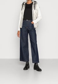 Stříbrná prošívaná vesta přes černé tričko, kombinovaná s tmavými džínami s širokými nohavicemi a černými robustními kotníkovými botami. Jednoduchý, funkční design.