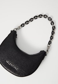 Borsa a spalla in pelle nera con forma curva, superficie testurizzata, logo argento e tracolla a catena con accenti neri e argento intrecciati.
