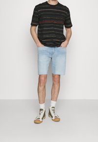 Man som bär svart kortärmad randig t-shirt, ljusblå denimshorts, vita strumpor och gröna och vita högstövlar, står med händerna i fickorna.