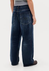 Person wearing dark blue wide-leg jeans with back pockets, one featuring a stitched oval logo, and low brown shoes against a plain background.