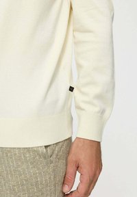 Világos krémszínű kötött pulóver bordás mandzsettákkal és szegéllyel. Oldalán egy kis fekete logó címke található. Bézs texturált nadrág felett viselve.