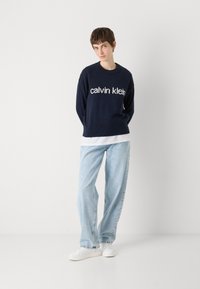 Jūrinė medvilnės megztinis su baltu "Calvin Klein" logotipu, derinamas su šviesiai mėlynomis laisvo kirpimo džinsais ir baltais sportbačiais. Paprastas, kasdieniškas apranga.