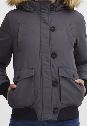 Veste matelassée gris foncé avec fermetures à boutons, poches à rabat devant, poignets côtelés et capuche bordée de fourrure, mains dans les poches.