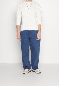 Sudadera blanca con cuello redondo, que presenta un pequeño logo. Combinada con unos vaqueros de denim azul holgados y zapatillas deportivas multicolor.