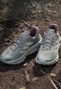 Scarpe da corsa grigio chiaro con tomaia in rete traspirante, dettagli bianchi e lacci rosa; progettate per l'uso all'aperto su terreni accidentati.