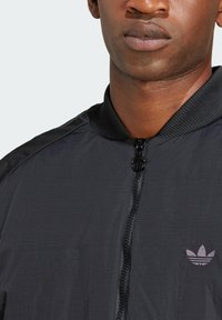 Casaco bomber leve preto com um acabamento texturizado, apresentando um fecho de zip e um pequeno logo bordado no lado esquerdo.