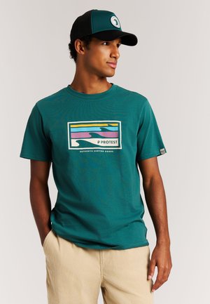 Turquoise katoenen T-shirt met een kleurrijke golfgrafiek en het woord "PROTEST." Gecombineerd met beige corduroy broek. Zwarte honkbalpet.