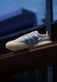 Vit sneaker med blå och vita ränder, gråmocka detaljer och en röd hälflik. Texturerad sula och slät yta. Höjd på en träytan.