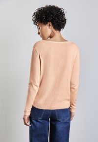 Pfirsichfarbener gestrickter Pullover mit weitem Rundhalsausschnitt und langen Ärmeln, kombiniert mit blauen Jeans. Weiche Textur und schlichtes Design.