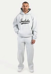 Sudadera con capucha de color gris claro con un logo negro, que incluye un bolsillo canguro y cordón ajustable, combinada con pantalones deportivos a juego en gris claro y zapatillas blancas.