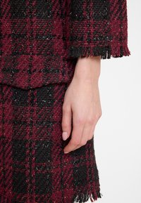 Rot-schwarzer kariert Stoff mit einer strukturierten Webart. Verfügt über fransige Kanten und ein glitzerndes Fadendetail, das eine taktile Oberfläche präsentiert.