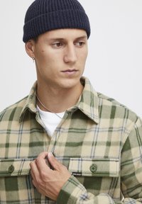 Geruite flanellen overhemd in groene en beige tinten, met twee voorzakken met groene knopen en een zachte textuur. Draagt een marineblauwe gebreide beanie.