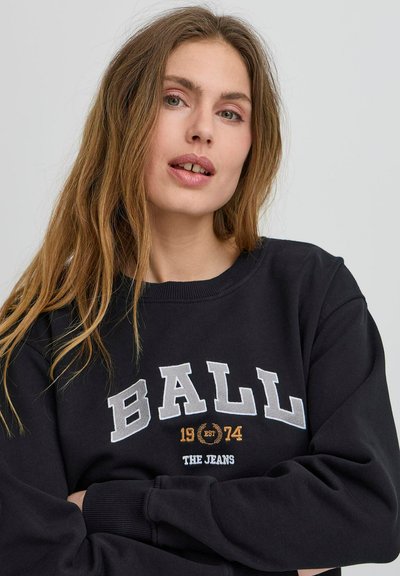 Fekete pulóver kerek nyakkivágással, fehér betűkkel a "BALL" szóval, arany színnel az "EST 74" felirattal, és alatta kisebb betűkkel a "THE JEANS" szöveggel.