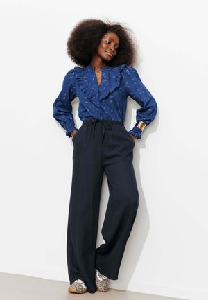 Femme aux cheveux bouclés volumineux portant un chemisier bleu à volants, un pantalon large noir, un bracelet en or et des chaussures à imprimé léopard, debout de manière détendue.